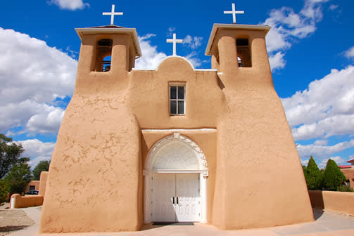 A bright adobe facade of a church with bright doors and crosses atop the steeples. A bright, partly cloudy sky is overhead.