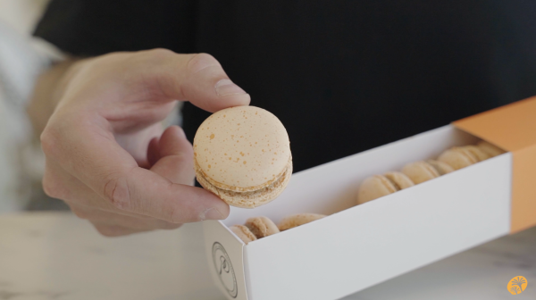 Image a sleeve of pumpkin spice flavored macarons with one pulled out and held towards to the camera for a closer look.