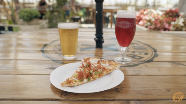 Slice of pizza on a wooden table, positioned in between two beers. One light, caramel colored beer to the left and one purple colored beer to the right.