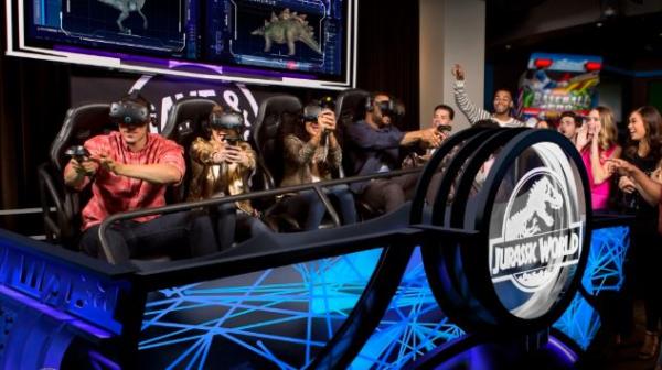 A group of adults wearing virtual reality goggles sitting on a ride with the Jurassic Park logo on the front at Dave & Buster's