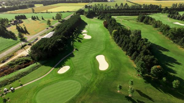 An aerial view of a golf course with wide green fairways, white sand bunkers, tree-lined edges, and farmland nearby.