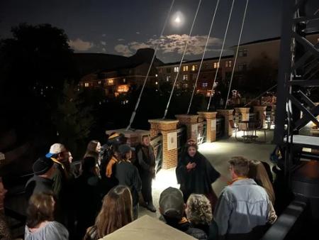 A female actor dressed in elegant period clothing tells a spooky story to a group during The Talking Dead Haunted Pub Crawl on the Washington Avenue Bridge in historic downtown Golden, Colorado. Castle Rock and a full moon loom in the background.