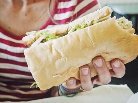 Woman wearing a pink and white stripped shirt holding a large sandwich in one hand.