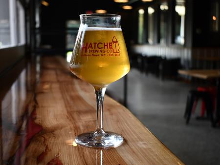 A glass full of Hatchet Brewing's craft beer with the red logo of Hatchet Brewing on the front of the glass sits on a wooden countertop.