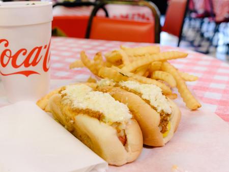 Two red hot dogs all the way laid out with fries, onion rings, and a Coke