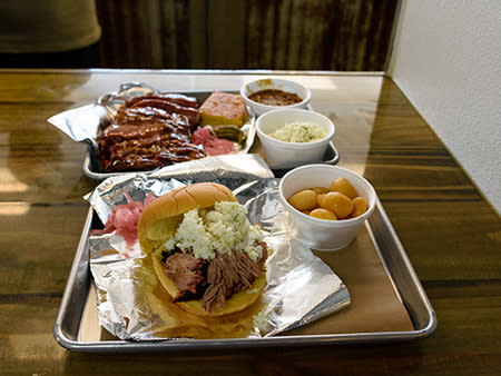 A BBQ pork sandwich sits on a metal tray with sides of tender pork strips, coleslaw, and baked beans.