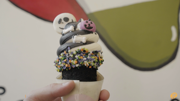 Image of a dessert being held. The dessert is a Chimney Cake that is coated in chocolate cookie crumbles and topped with a black and white soft-serve. The toppings include Halloween sprinkles and decorations.