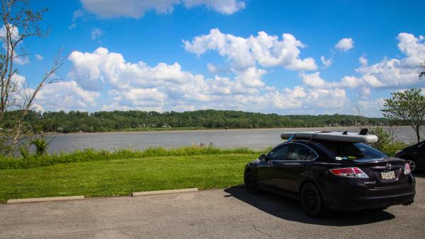 Car with kayaks secured to the roof parked beside the Ohio River at Charlestown State Park on a sunny day with blue skies and clouds.