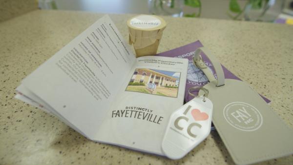 A close up of the DistiNCtly Fayetteville Tourism Passport on a counter alongside a luggage tag, an “I ❤️ CC” keychain, and a small branded gift from the visitors center.