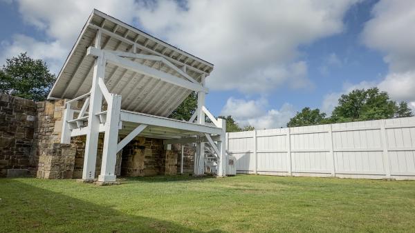 A photo of the white wooden gallows at Fort Smith National Historic Site shot in front of a cloudy blue sky.