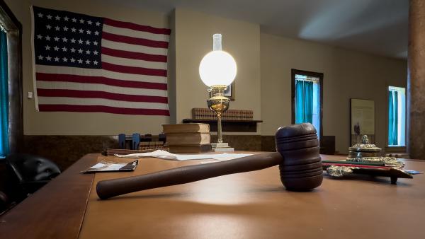 A gavel placed on a desk is backlit by an antiquated lamp while an American Flag hangs on the wall.