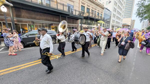 A male brass band plays musical instruments while people with decorated umbrellas dance in the street