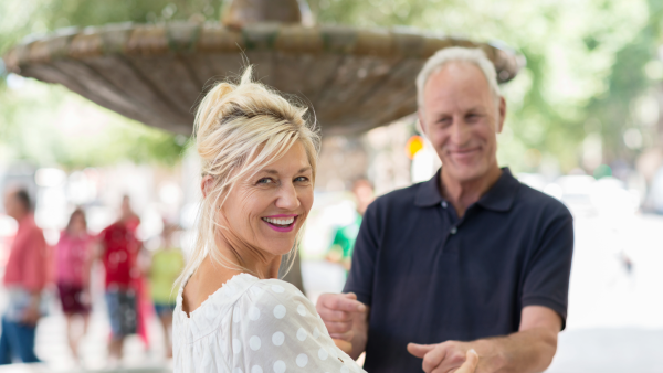 A man in a blue polo shirt dances with a blond woman in a white blouse in front of a fountain