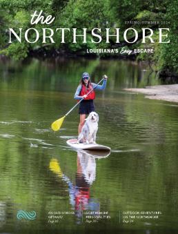 The cover of The Northshore: Louisiana's Easy Escape Visitor Guide, Spring-Summer 2026 issue, which features a woman and her dog atop a paddleboard on the Bogue Falaya River in Covington. She is using a yellow paddle to navigate.