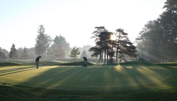Two golfers putting on a green golf course during sunrise, with long shadows and misty trees in the background.