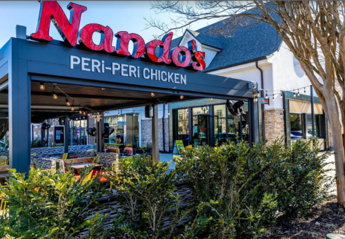 Exterior seating area of a restaurant with a large red sign that says "Nando's". Large shrubs in the front.