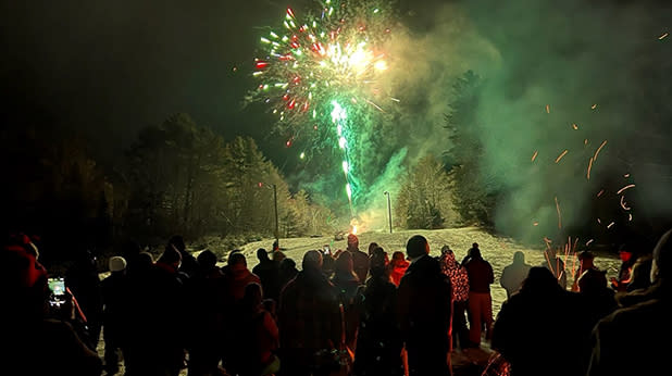 A crowd of people stands on snow-covered ground, watching vibrant fireworks bursting in the night sky at the Long Lake Winter Carnival.