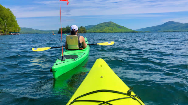 POV shot from a yellow kayak while a person in a green kayak paddles on Lake George with a view of the mountains in the background