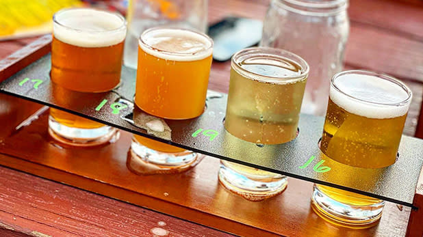Four beer-tasting glasses in golden, amber, and pale hues sit on a wooden flight board, creating a cheerful, inviting bar scene at Hunter Mountain Brewery.