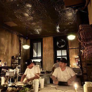 Two pizza makers in white t-shirts working behind the prep counter of a dimly lit, rustic New York pizzeria with exposed brick walls, a tin ceiling, and pendant lighting.