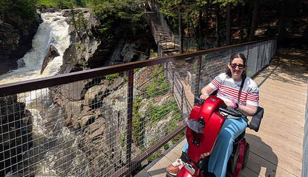 A person in sunglasses smiles on a red mobility scooter on a wooden bridge at High Falls Gorge. A waterfall cascades beside them, surrounded by lush greenery.