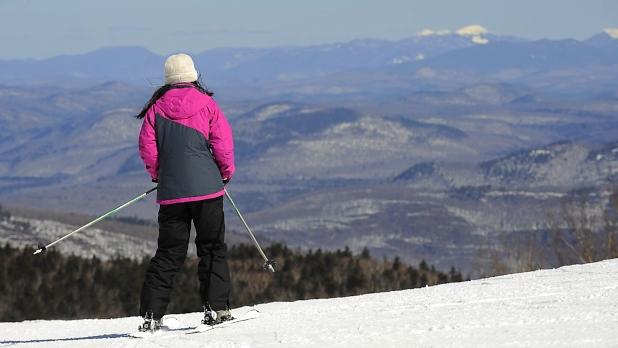 A person in a pink and grey winter coat, white hate, and black snow pants prepares to ski down Gore Mountain