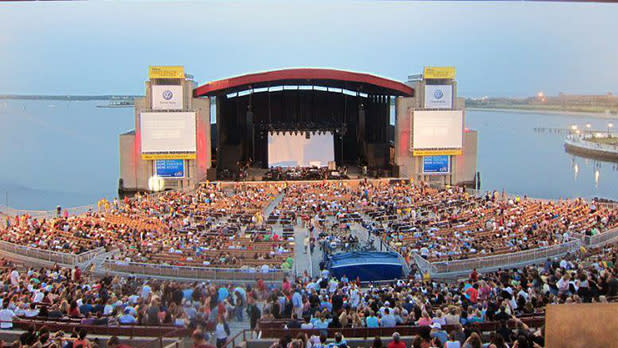 People sit to enjoy a performance at an open air amphitheater with waterfront views at Northwell Health at Jones Beach Theater