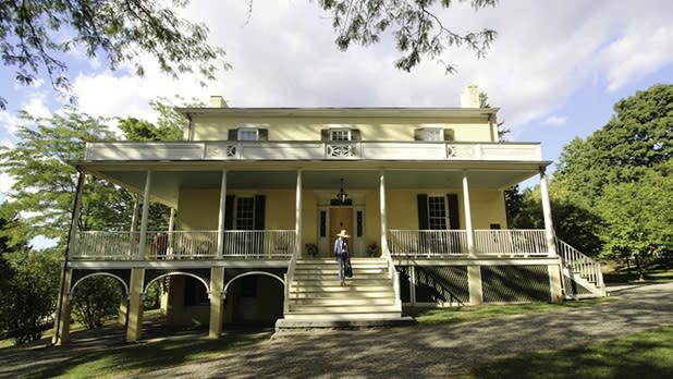 A person in a sun hat walks up the steps of a yellow house at the Thomas Cole National Historic Site