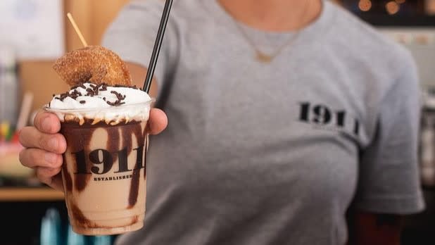 A person holding a chocolate drink topped with chocolate sauce, whipped cream, and a cider donut