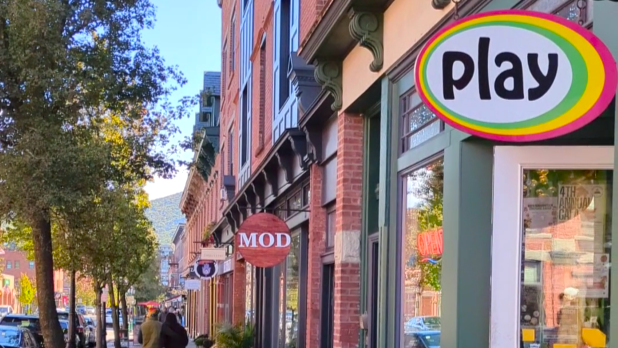 Beacon's Main Street lined with brick buildings and colorful shop signs, including "play" and "MOD." Trees line the sidewalk under a clear blue sky.