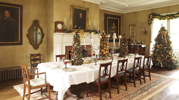 Opulent dining room with festive decor. A large table is set with a white cloth, surrounded by chairs. Christmas trees and garlands enhance the cozy elegance at Locust Grove Estate in Poughkeepsie, New York.