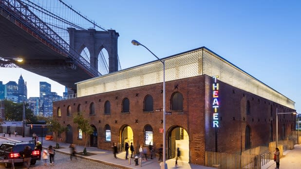 A neon rainbow "theater" sign on the brick exterior of St. Ann's Warehouse with the Brooklyn Bridge in the background