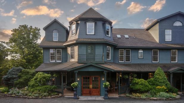A charming, large blue Victorian house with ornate details is surrounded by lush greenery under a sky with scattered clouds, creating a serene atmosphere.