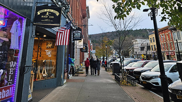 Shops and cars line Main Street in Cooperstown with lush green hills in the background