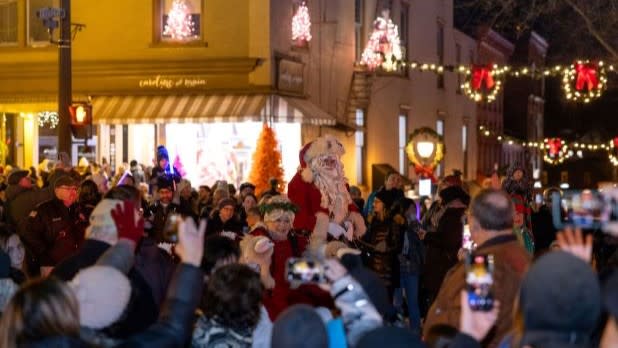 A festive street scene with Santa Claus walking through a cheerful crowd at night at the Saratoga Victorian Streetwalk. The street is adorned with twinkling lights and holiday decorations.