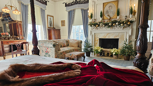 Bedroom within Westbury House at Old Westbury Gardens with elegant furnishings, a canopy bed, floral sofa, ornate fireplace adorned with garland, and festive decor.