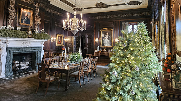 Elegant dining room with dark wood panels, lit chandelier, and ornate fireplace. A decorated Christmas tree adds a festive, cozy atmosphere during Christmas at Westbury House at Old Westbury Garden.