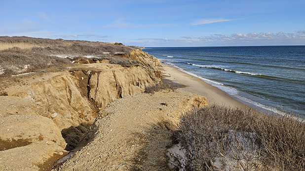 Coastal scene at Shadmoor State Park with eroded sandy cliffs overlooking a narrow beach and calm ocean. The sky is clear, suggesting a serene and peaceful setting.