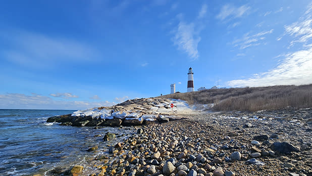 A picturesque shoreline with a rocky beach leading to the Montauk Point Lighthouse in the distance. The sky is blue with scattered clouds, creating a serene and tranquil scene.
