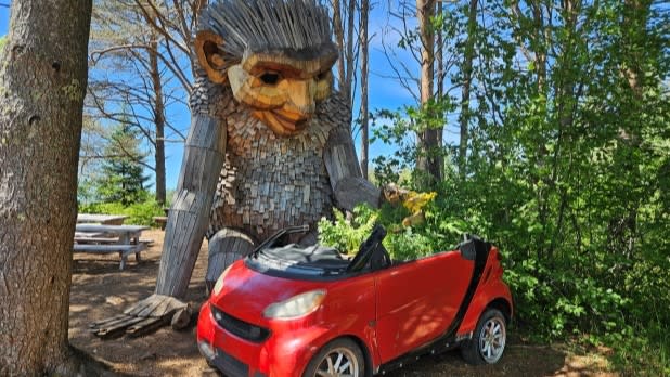 A large wooden sculpture of a friendly, mythical creature kneels in the woods by a small red convertible during Thomas Dambo's Trolls exhibit at the Wild Center in Tupper Lake.