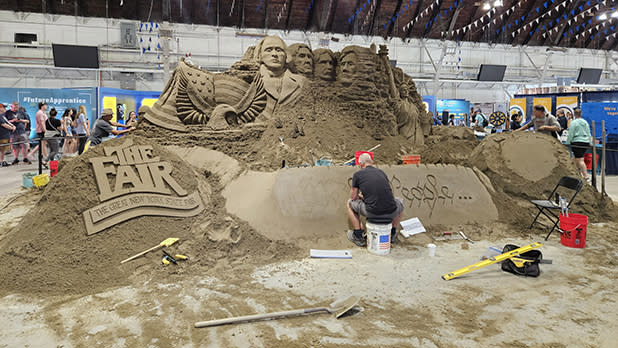 A detailed sand sculpture featuring a replica of Mount Rushmore and bold lettering "THE FAIR" above a banner reading "THE GREAT NEW YORK STATE FAIR."