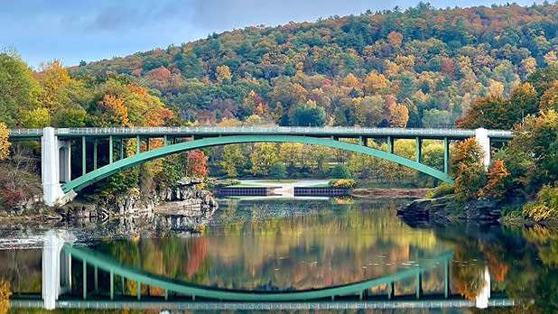 A green bridge spans a waterway in Narrowsburg with trees decked in fall colors in the background