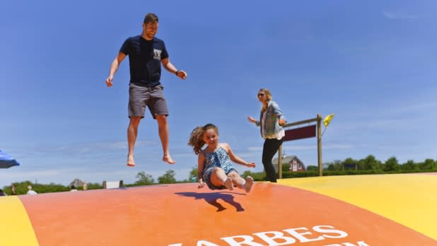 Adult man and two children jumping on an oversized trampoline-like bounce pillow; clear blue sky in the background