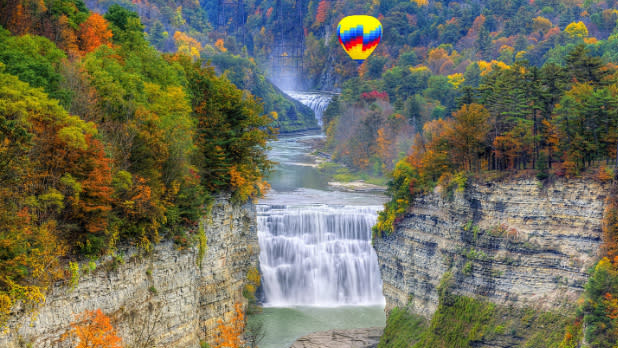 A yellow hot air balloon with blue and red stripes flying over Letchworth Falls in Fall