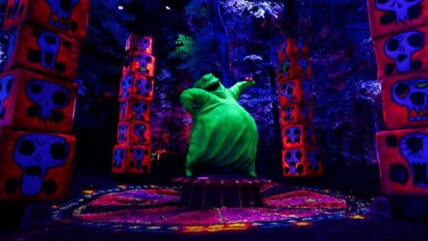 A large green character stands on a platform surrounded by neon-lit blocks with skull designs in a dark, colorful environment at The Nightmare Before Christmas Light Trail at New York Botanical Garden.