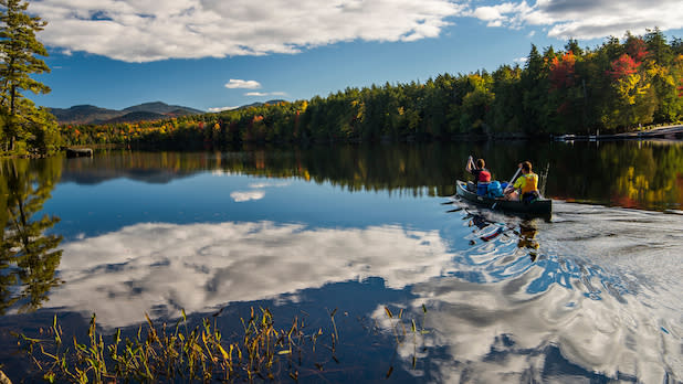 Two people paddle in a canoe on the Saranac Lake Chain with a view of early fall colors throughout the Adirondack Mountains