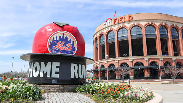 Large red apple with a Mets logo and "Home Run" sits near Citi Field, NYC. Tulips bloom in front, creating a vibrant, lively atmosphere.
