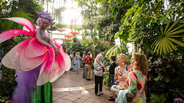 A performer in a vibrant flower costume with pink petals interacts with smiling guests in a lush conservatory, creating a whimsical, joyful atmosphere at the Buffalo and Erie County Botanical Gardens