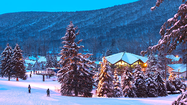 A group of people walking along the snow in front of Bristol Mountain ski resort in the evening. Purple lights are reflected on the snow, large pine trees and glowing lights can be seen from inside the glass walls of the resort.