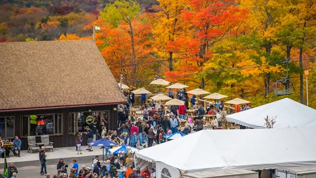 A crowd of people mingling between various tents surrounded by the oranges, reds, and, yellows of fall